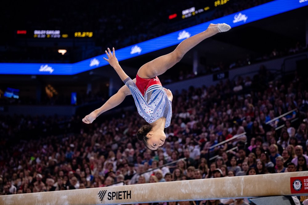 A gymnast does a flip on a balance beam while wearing a red, white and blue leotard.
