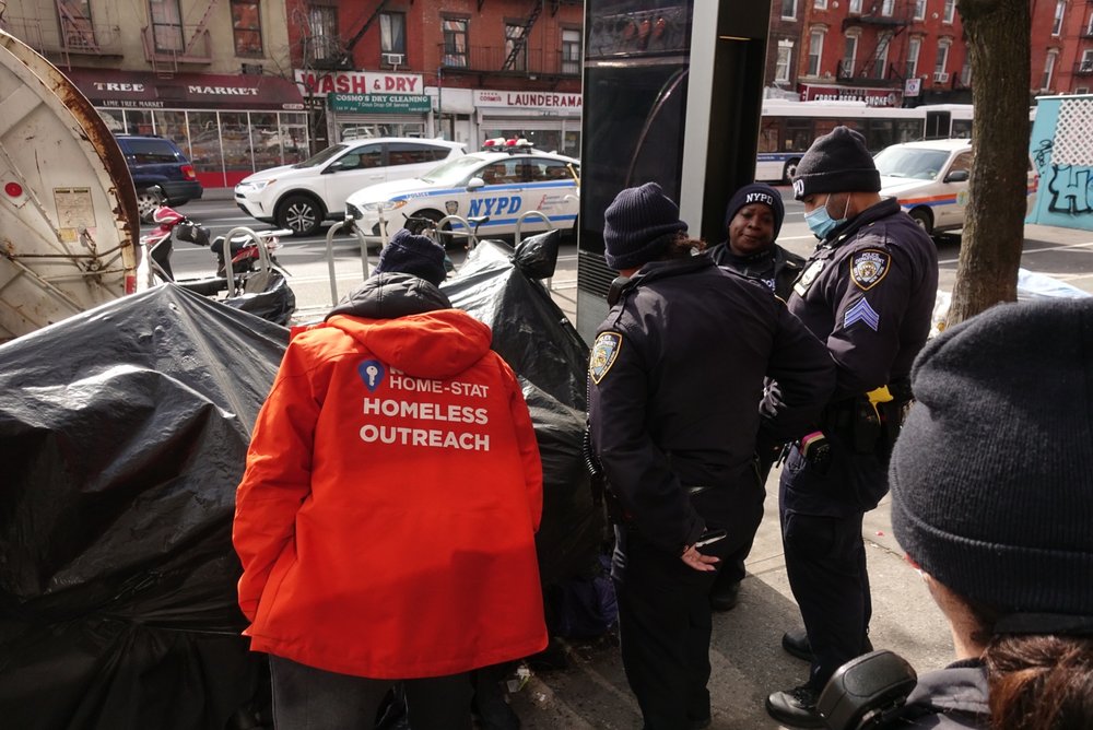 Police and a person wearing a red jacket saying "homeless outreach" surround a homeless encampment