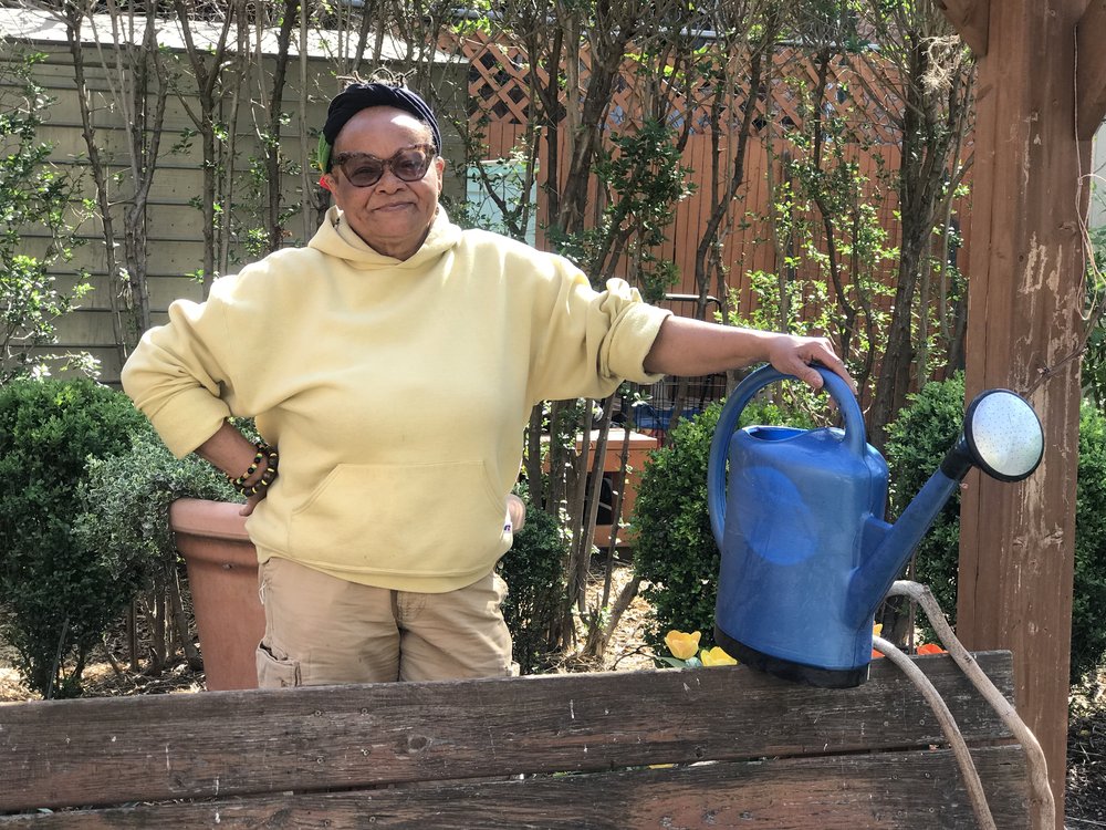 community gardener Ena K. McPherson standing with a blue gardening water pitcher in one hand. She is wearing a yellow sweathshirt and hat