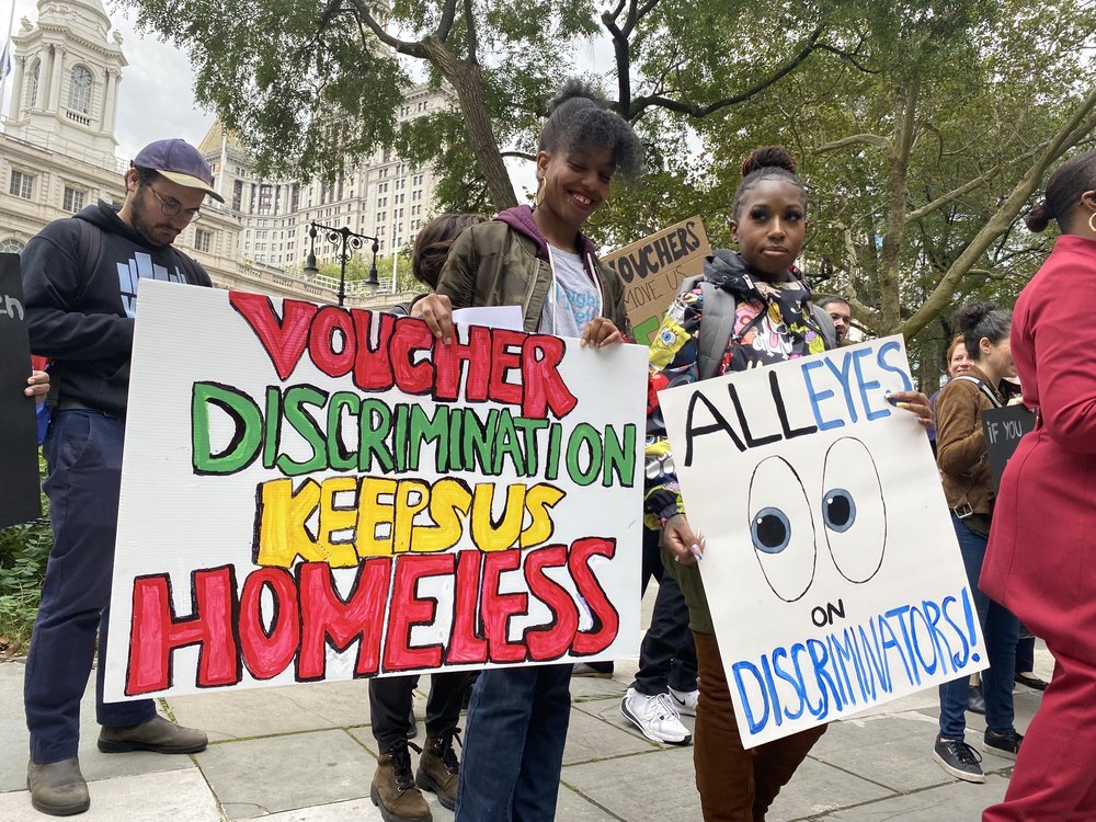 Outside of City Hall, advocates hold colorful signs, including one that says "voucher discrimination keeps us homeless."