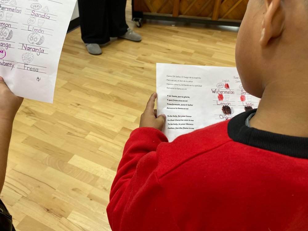 A young boy in a red sweatshirt clutches a worksheet with lyrics in english and spanish.