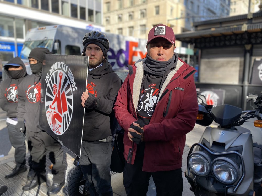 Two men at a press conference near City Hall hold a sign of a man on a bike, the logo for Los Deliveristas Unidos.