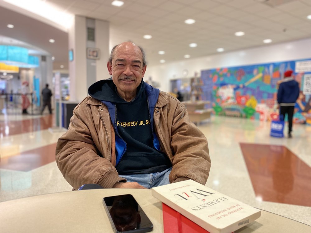 Philip Malebranche, 65, sits at a table, wearing a sweatshirt with a tan jacket over it.