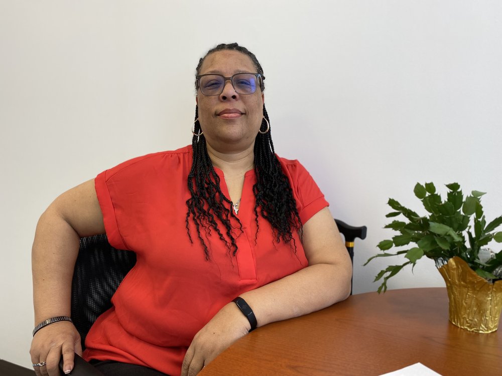 Cynthia English sits leaning on a table, wearing a red shirt and braids in her hair.