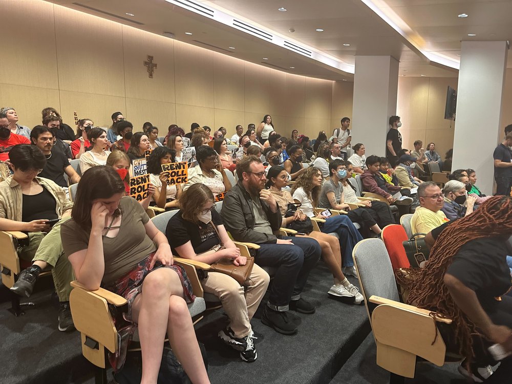 A crowd of people sit in an auditorium, some with orange "rent roll back" signs.