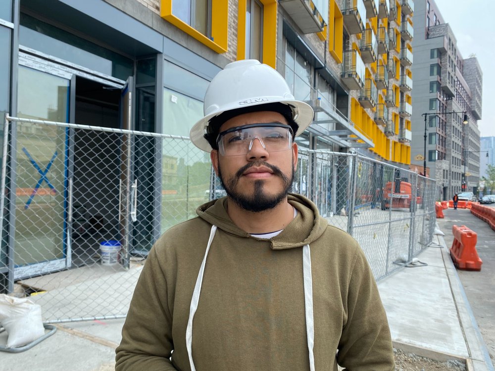 Construction foreman Anthony Santillan poses for a picture in front of a fenced building with a white hard hat on.