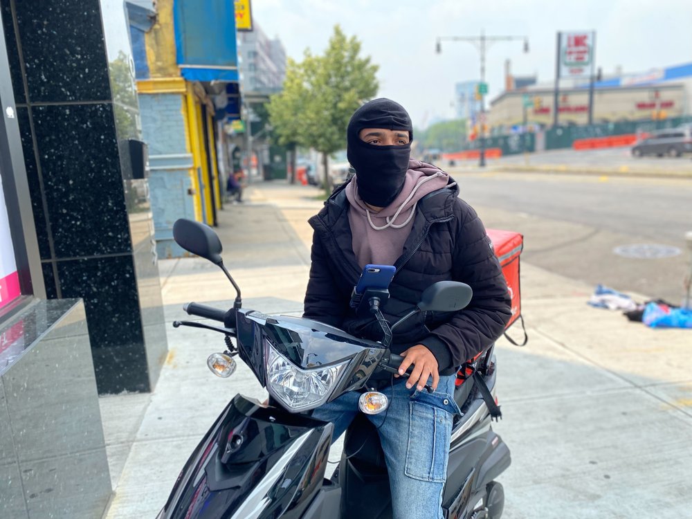 App-based food delivery worker Anthony Santillan sits on his motorcycle with a black face balaclava covering his face.