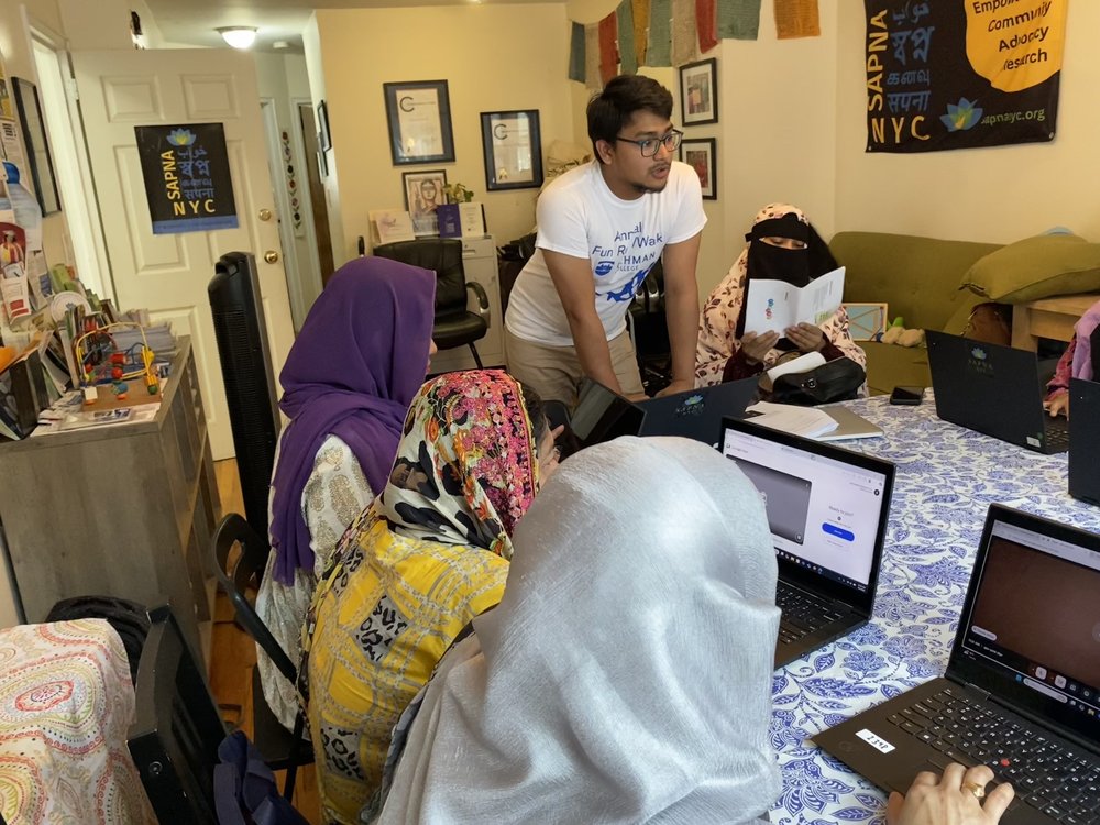 Three women wearing colorful headscarves sit at laptops during a computer literacy class in the Parkchester neighborhood of the Bronx, while a male instructor in glasses and a white t-shirt stands nearby providing guidance. The room features educational posters and certificates on the walls.