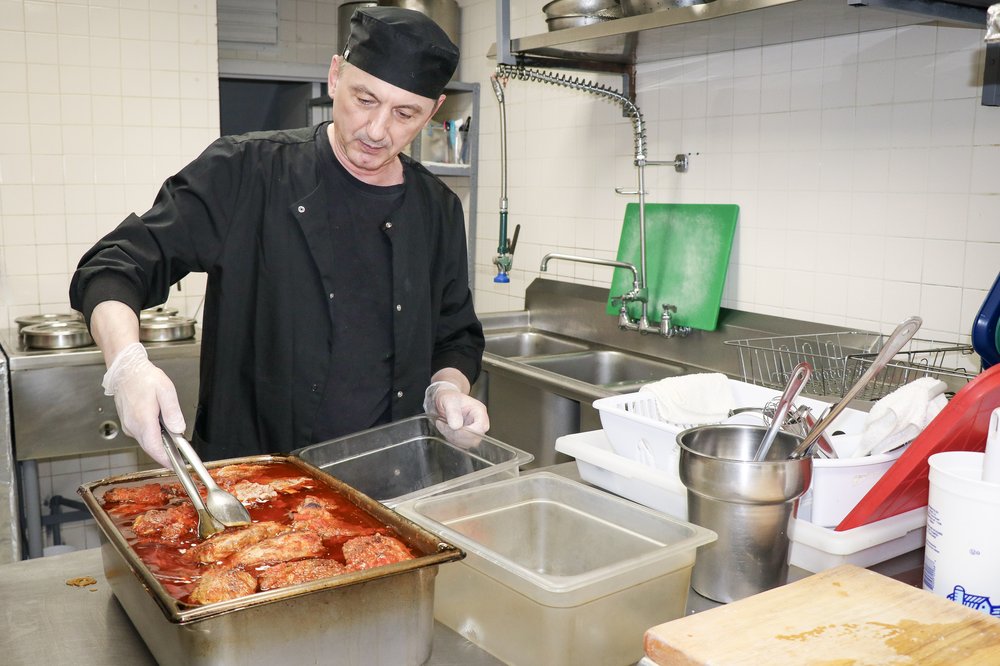 A chef works in a kitchen using kitchen tongs to move items from a silver dish.