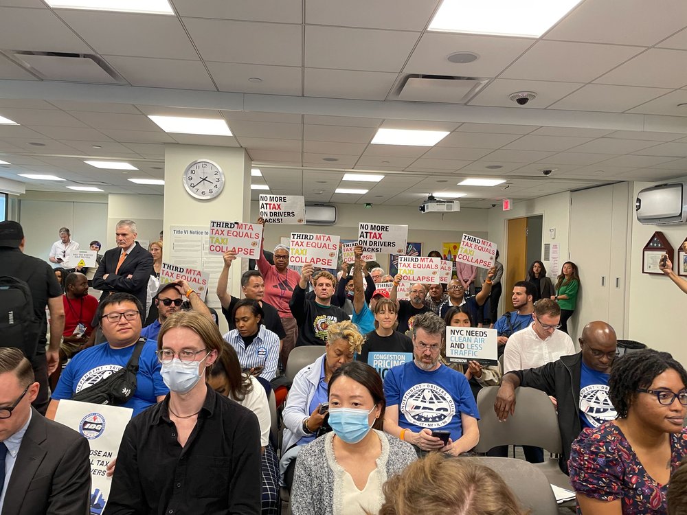 Dozens of people, some carrying signs against a toll, sit waiting for the MTA meeting to begin.