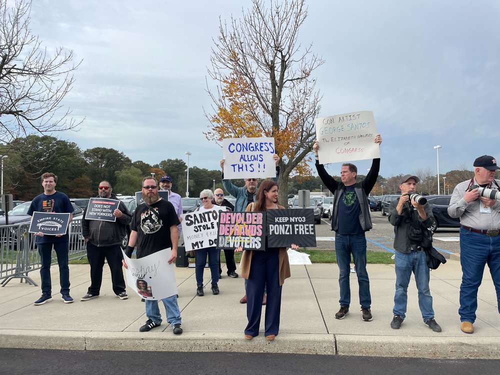 Demonstrators hold signs outside of the court house where Santos is being arraigned. One sign says "Devolder defrauds devoters."
