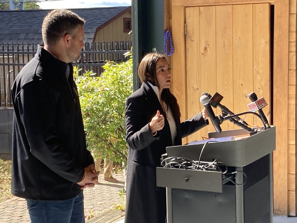 Rep. Alexandria Ocasio-Cortez speaks at a podium beside Rep. Pat Ryan.