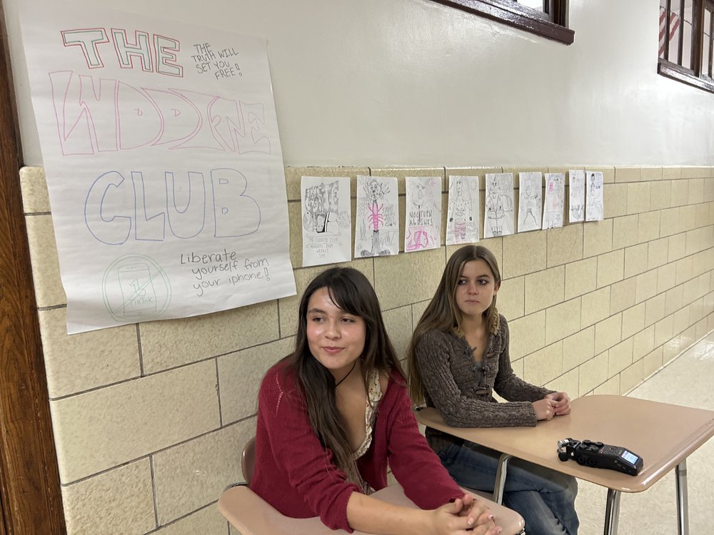 Two girls sit at desks beneath a "Luddite club" poster.