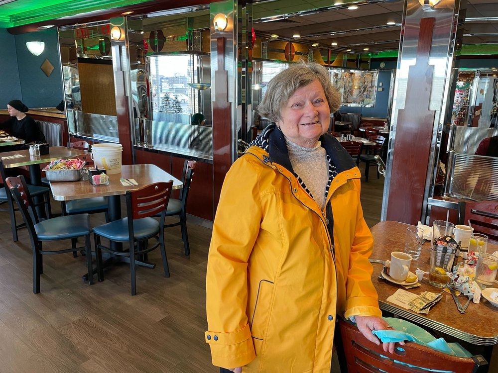 Maryellen Conway, in a yellow raincoat, stands by a table at the Carle Place Diner.