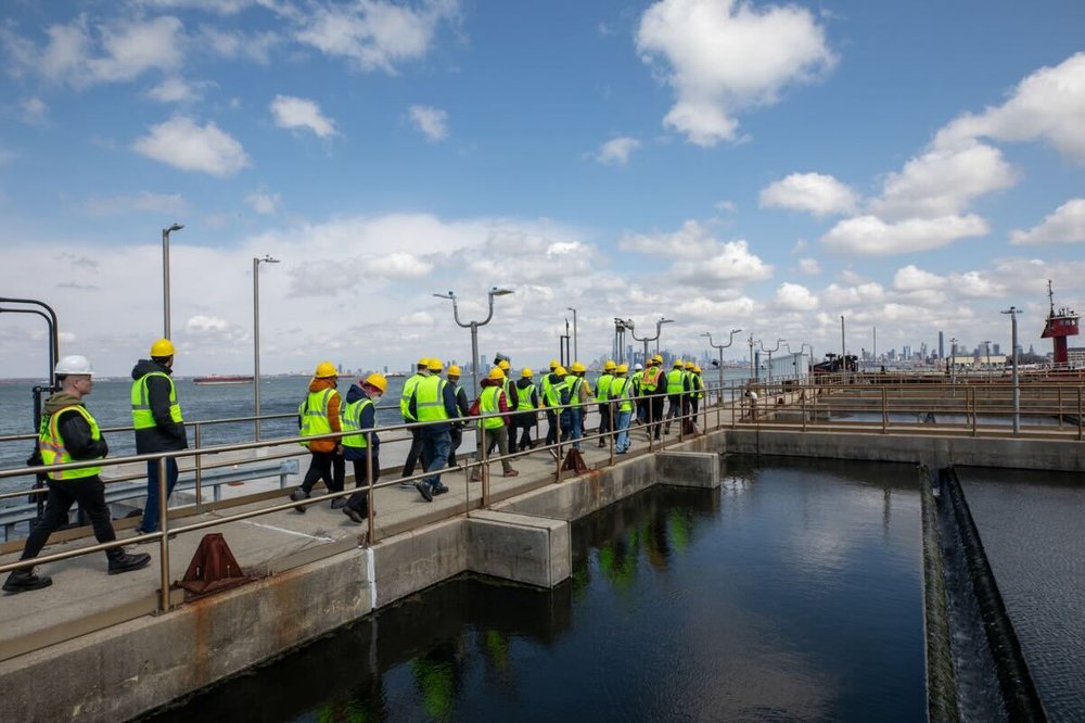 A group of people in yellow hard hats and vests line the walkway near the water.