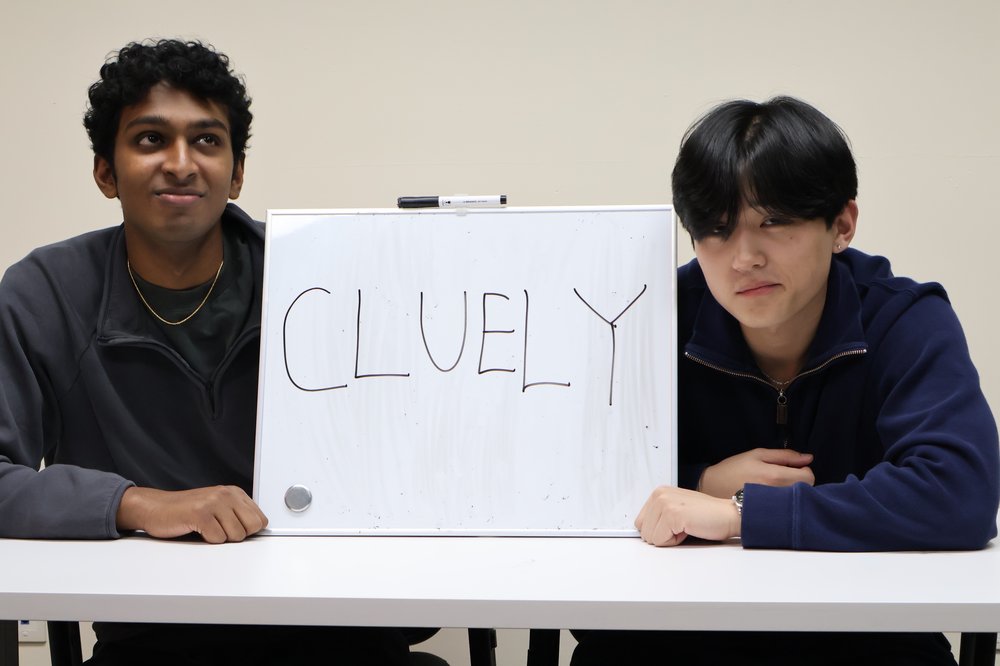 Two people sit at a table holding a sign that says "Cluely."