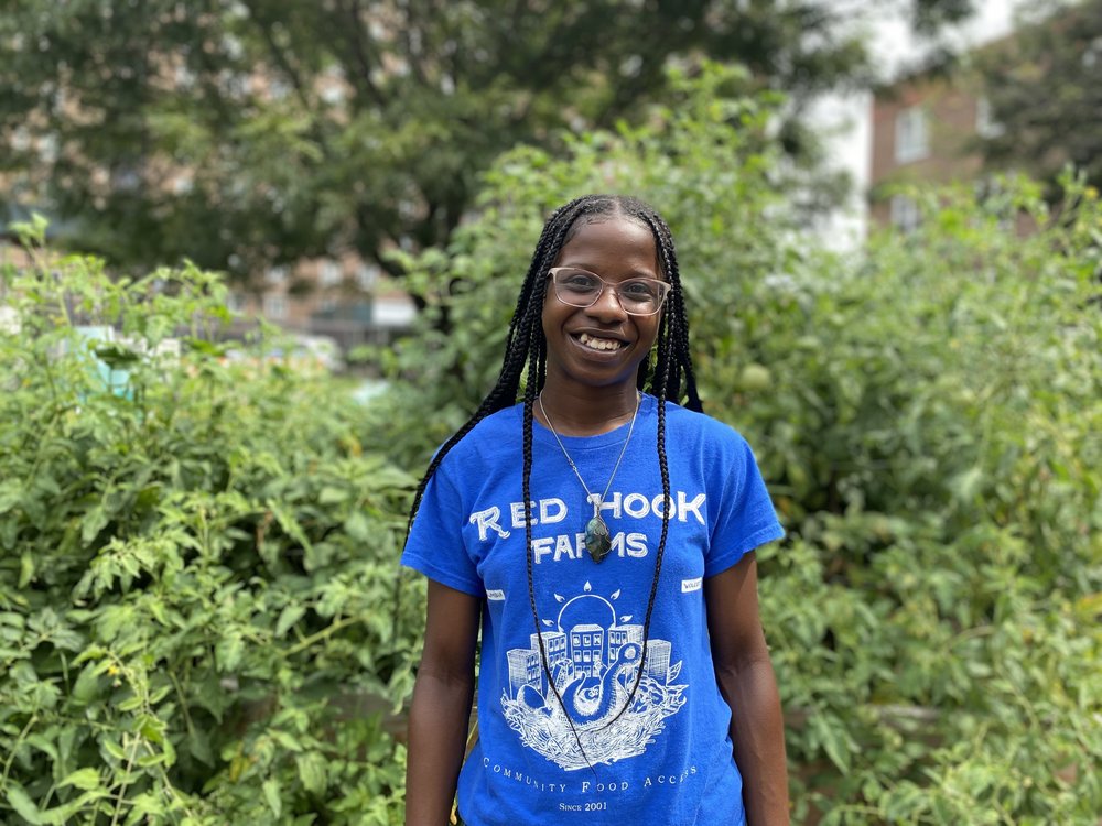 Wolcott Farm Coordinator Chane Wright, stands in front of plants while wearing a blue shirt that says Red Hook Farms.