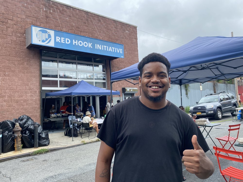 Quincy Phillips flashes a smile and a thumbs up as he stands in front of a building with a blue awning that says Red Hook Initiative.
