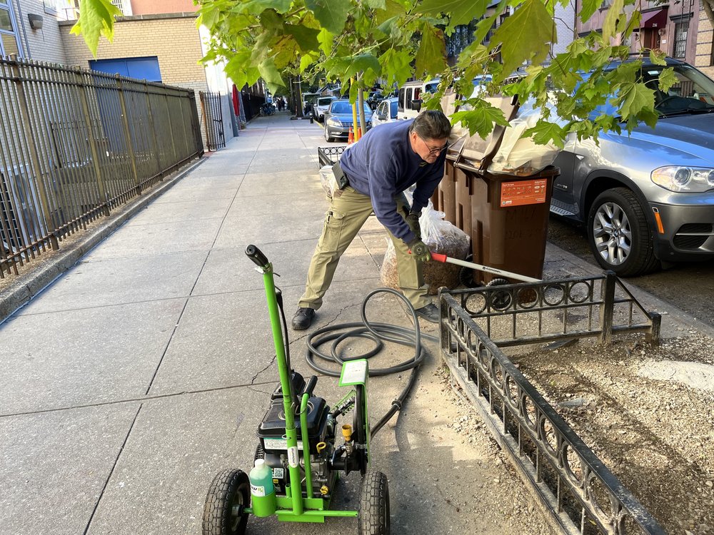 An exterminator on a sidewalk with a green machine that looks similar to a lawnmower with a hose attached.