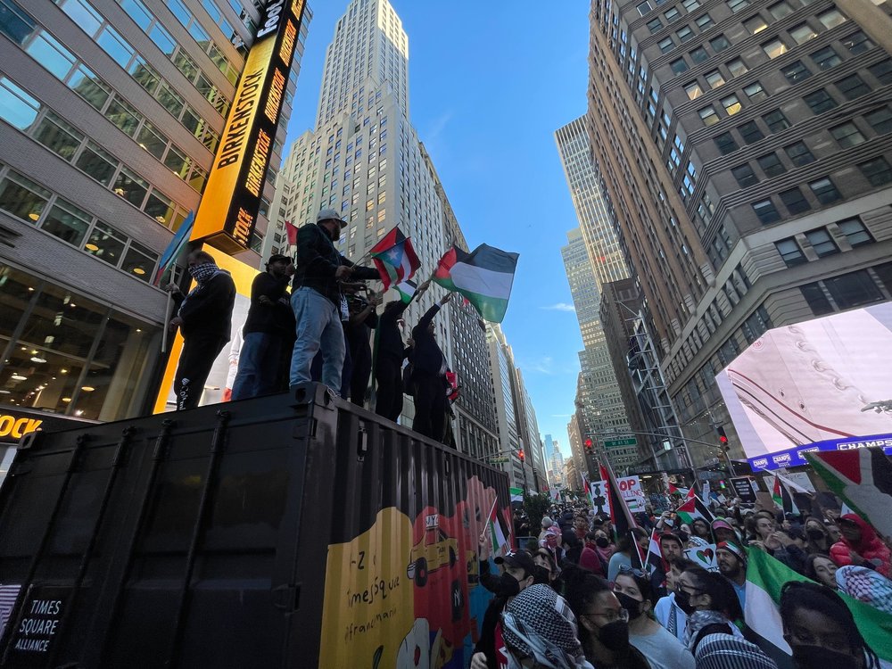 Crowds of people stand on a shipping container and wave Palestinian flags.