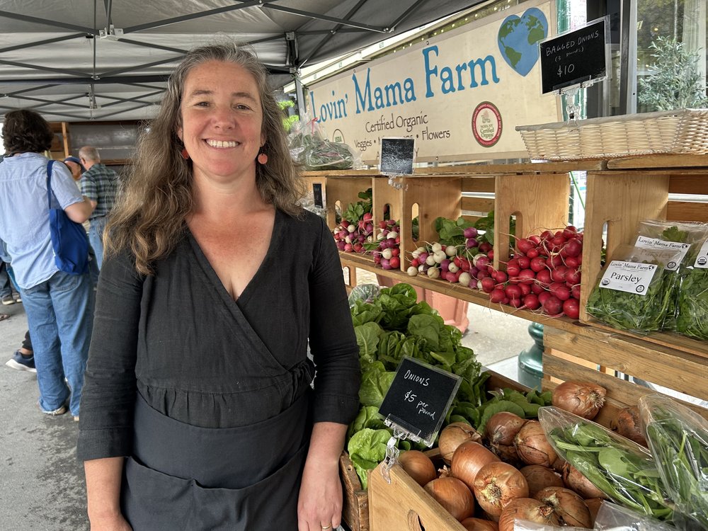 A woman smiles in a stand at a farmers' market