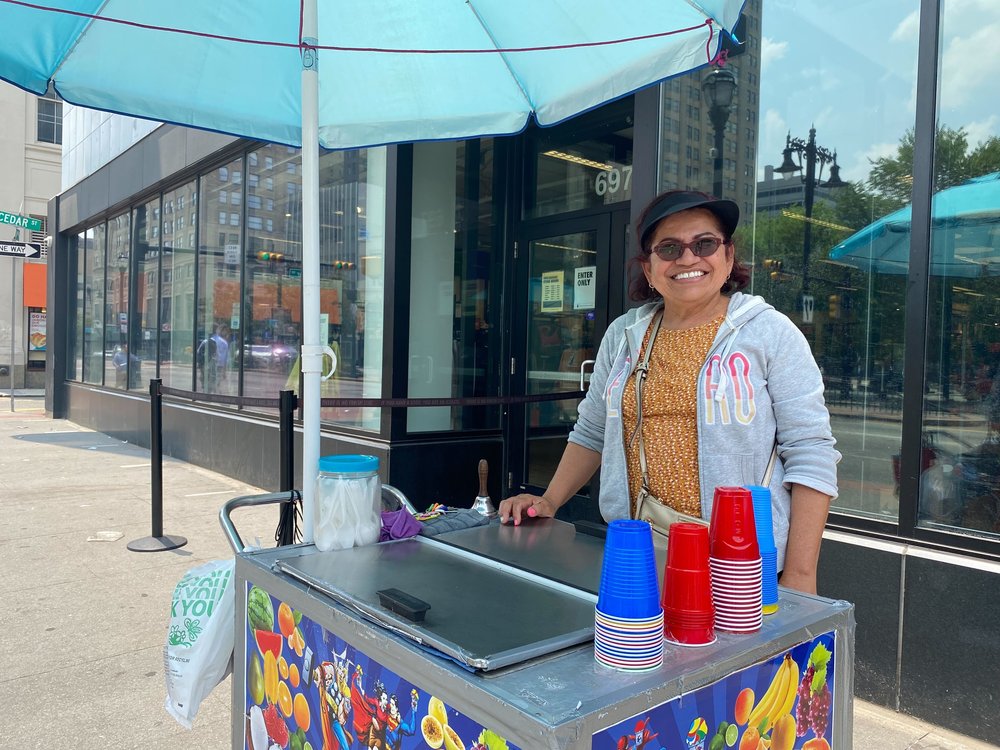 Ice cream vendor Carmen Rojas wears a visor, sunglasses as an umbrella shades her and her trolly.