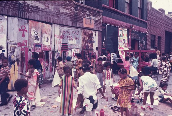 A vintage photograph of  a large group of children milling about on the sidewalk in front of a community center.