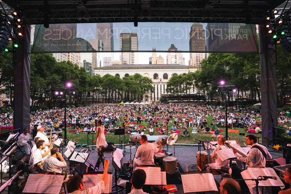 A band onstage, seen from behind, with an audience sprawled out before them on a lawn