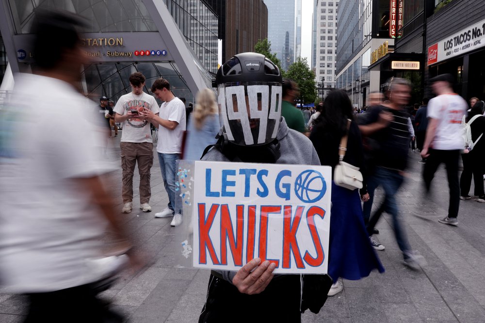 A photo of a Knicks fan in a motorcycle helmet with the year 1999 written across the front, while holding a homemade Go Knicks sign in orange and blue.