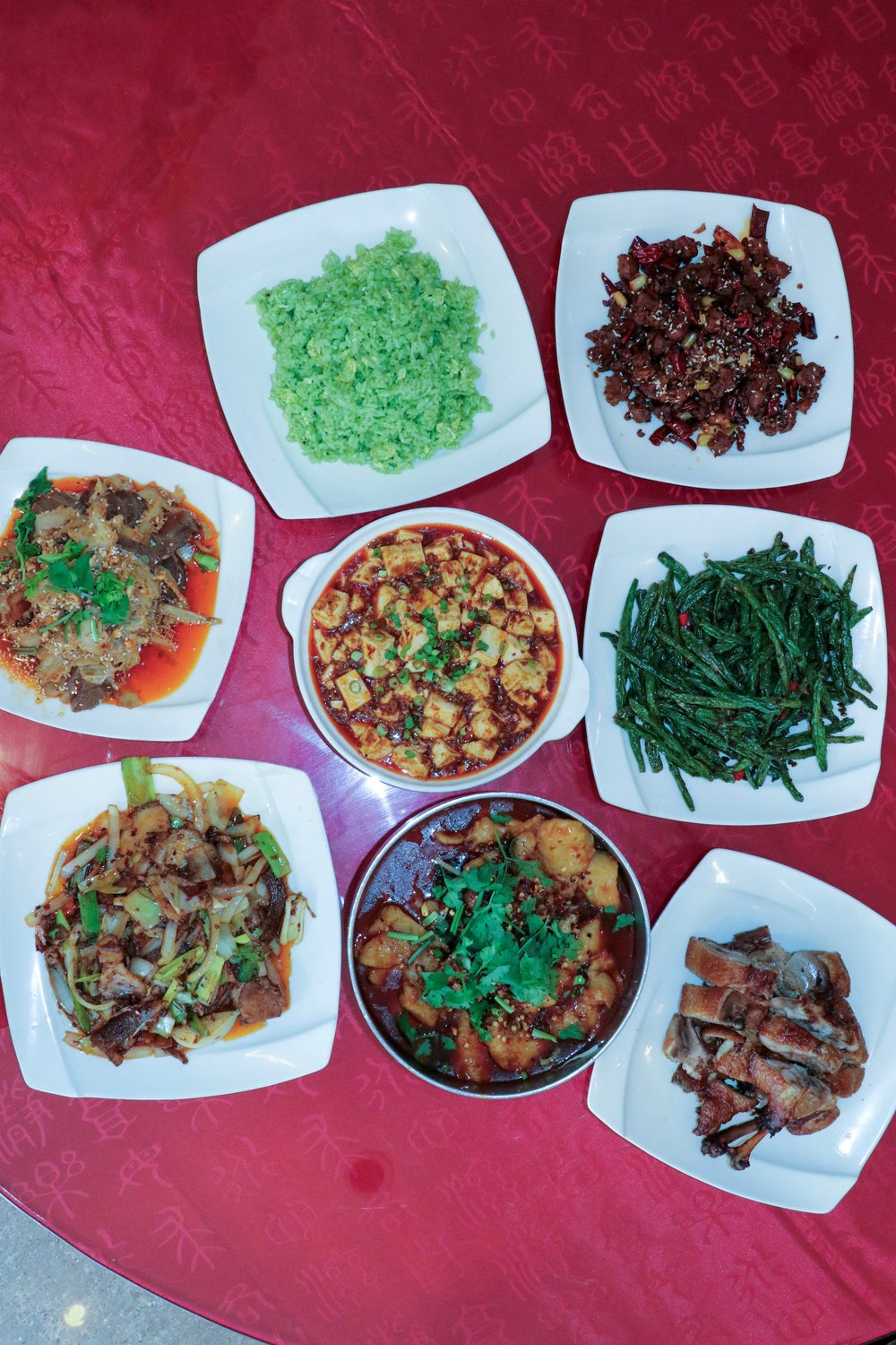 An array of 8 dishes of Sichuan food on a red table.