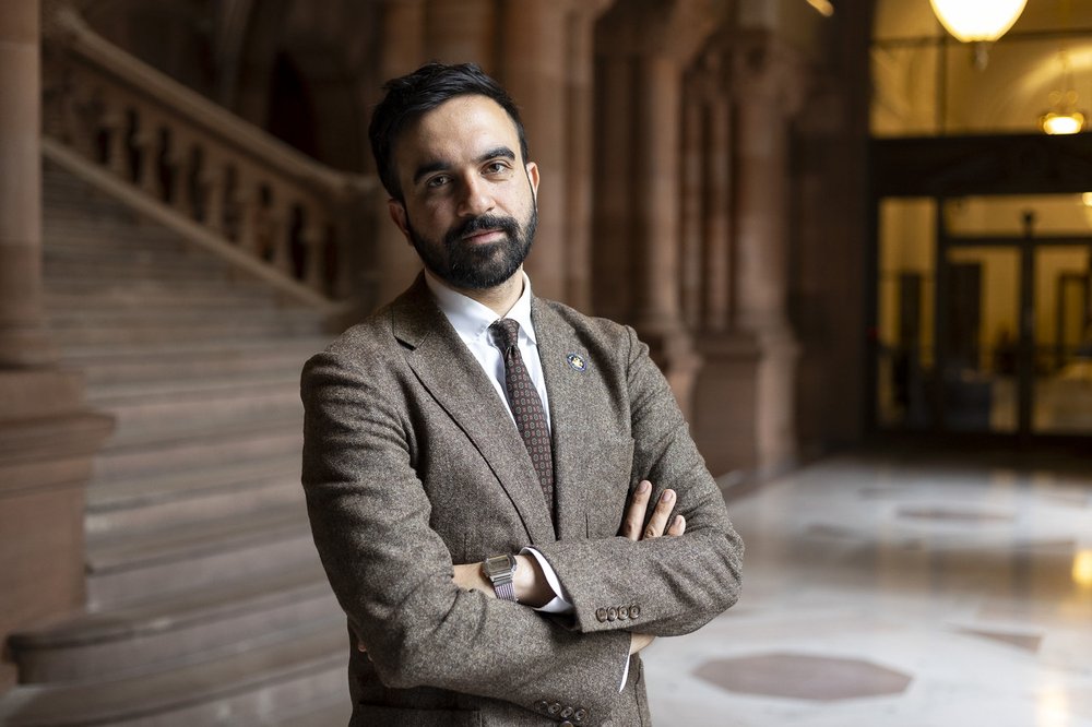 Zohran Mamdani in a suit with his arms crossed.