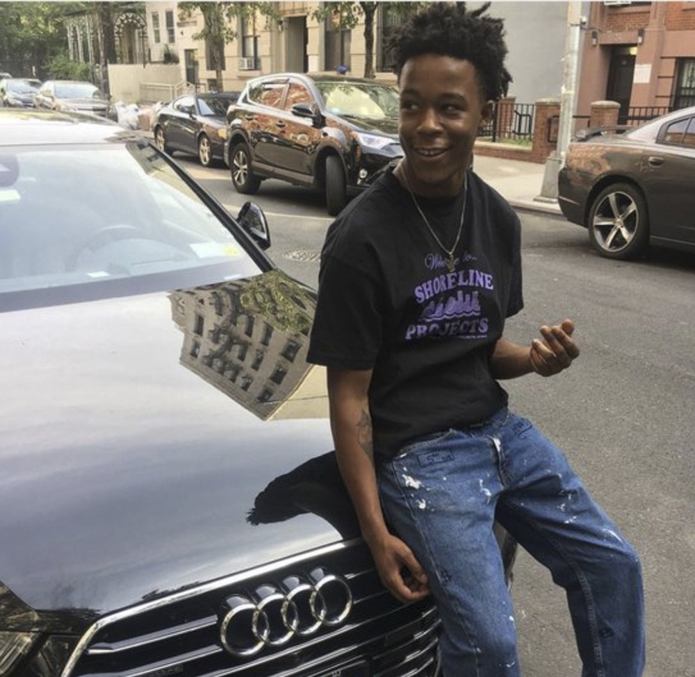 A smiling young man sits on the hood of a car.