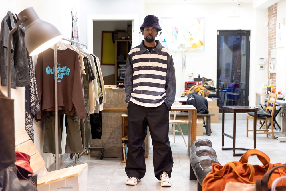A man in a striped sweater and bucket hat stands in the middle of a clothing store.
