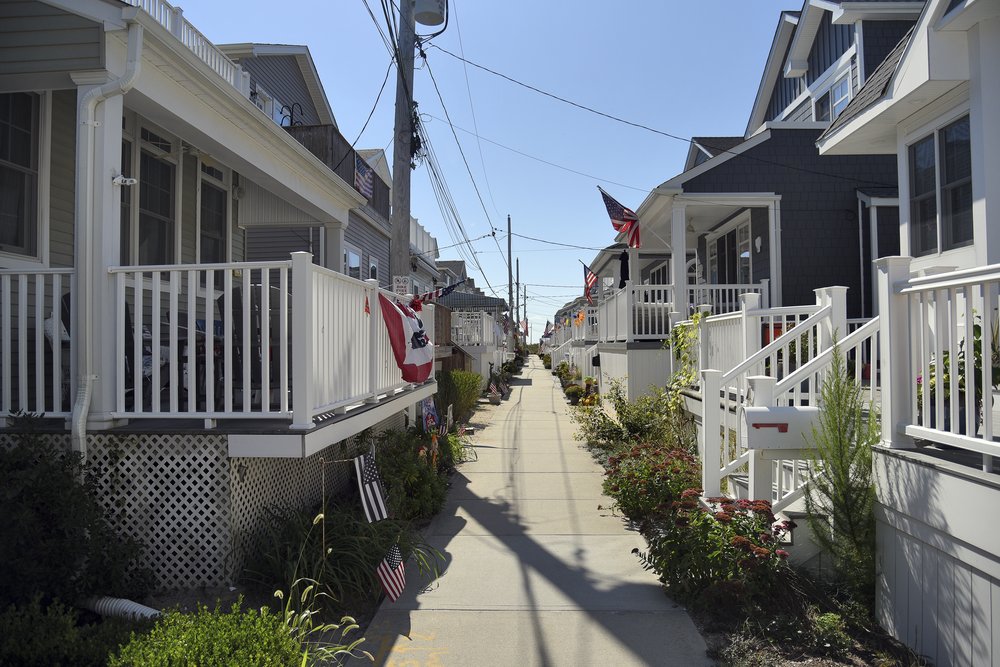 An alleyway between homes on a sunny day. American flags adorn the porches of most of the homes, which face each other.