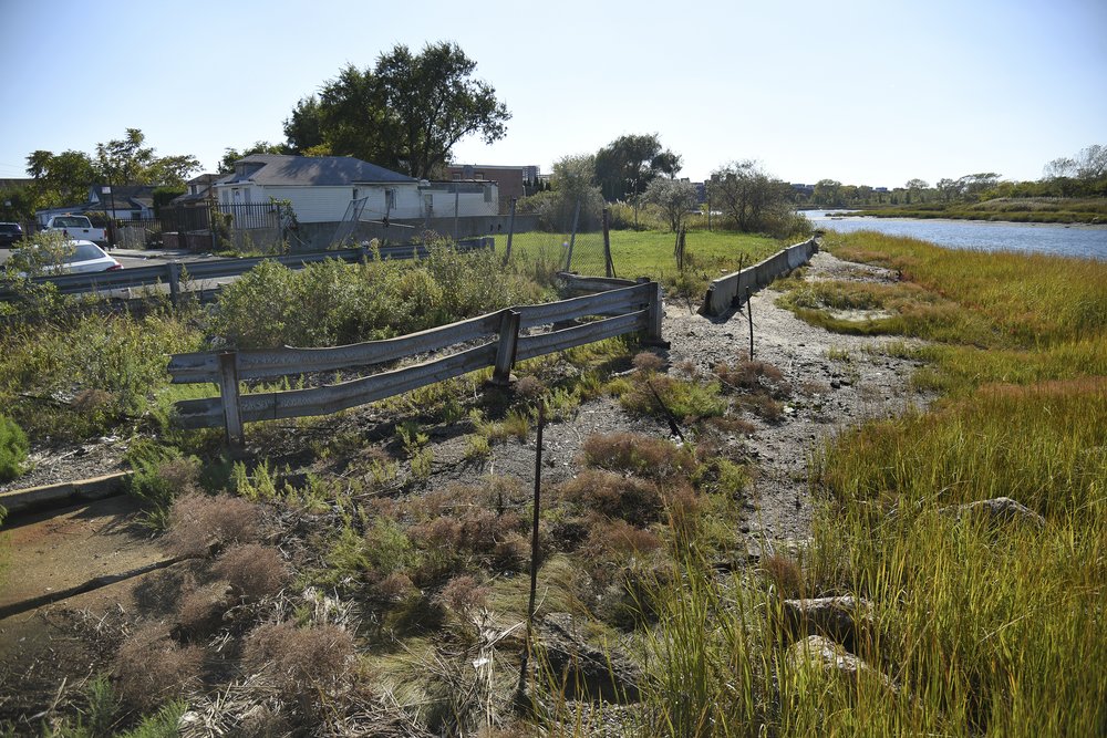 Houses along the bayside of Edgemere, a neighborhood in Rockaway, Queens. Grass, dirt, and a broken fence fill the frame. he neighborhood, which has been neglected by the city for decades, has seen no substantial infrastructural development to protect against the next storm.