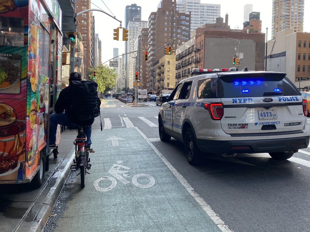 A photo of a biker waiting next to a police car at a traffic light.