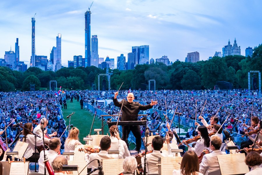 A conductor leading an orchestra in an outdoor concert with an audience in view.