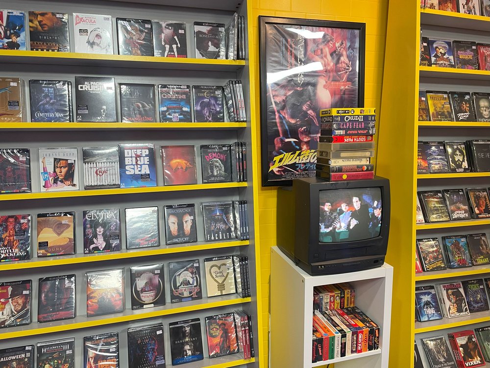 The interior of a video rental store with movies lined up against a yellow wall.