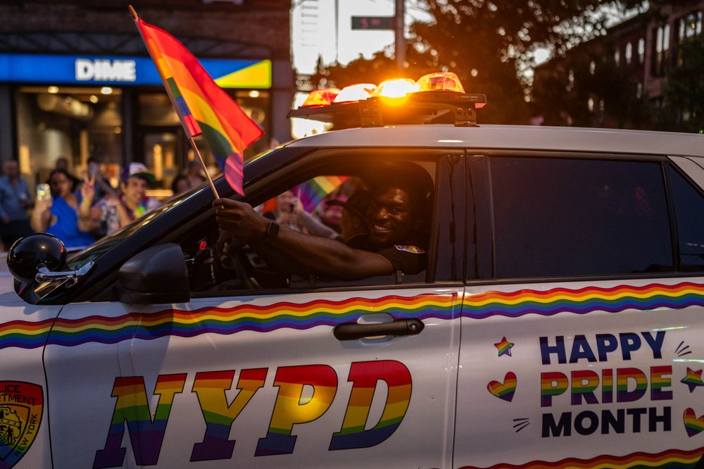 A photo of a police officer smiling while driving an NYPD cruiser decked out in rainbow colors.