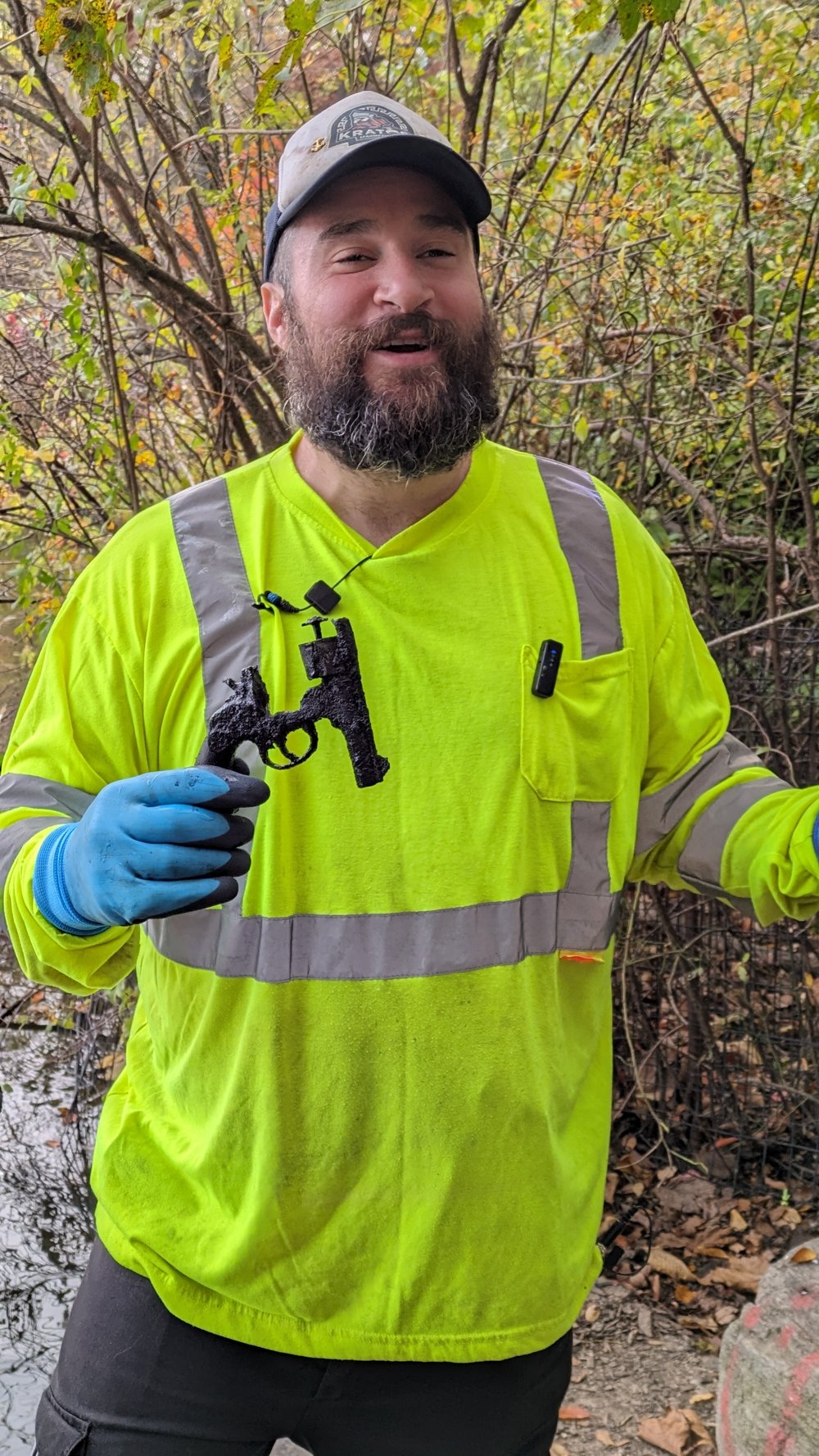 A bearded man in a yellow construction jacket and cap smiles and holds a rusty revolver.
