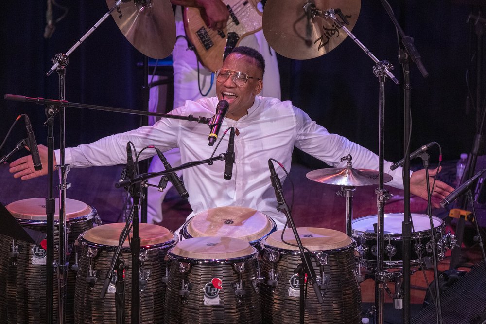 A percussionist dressed in white, singing behind a large array of drums.