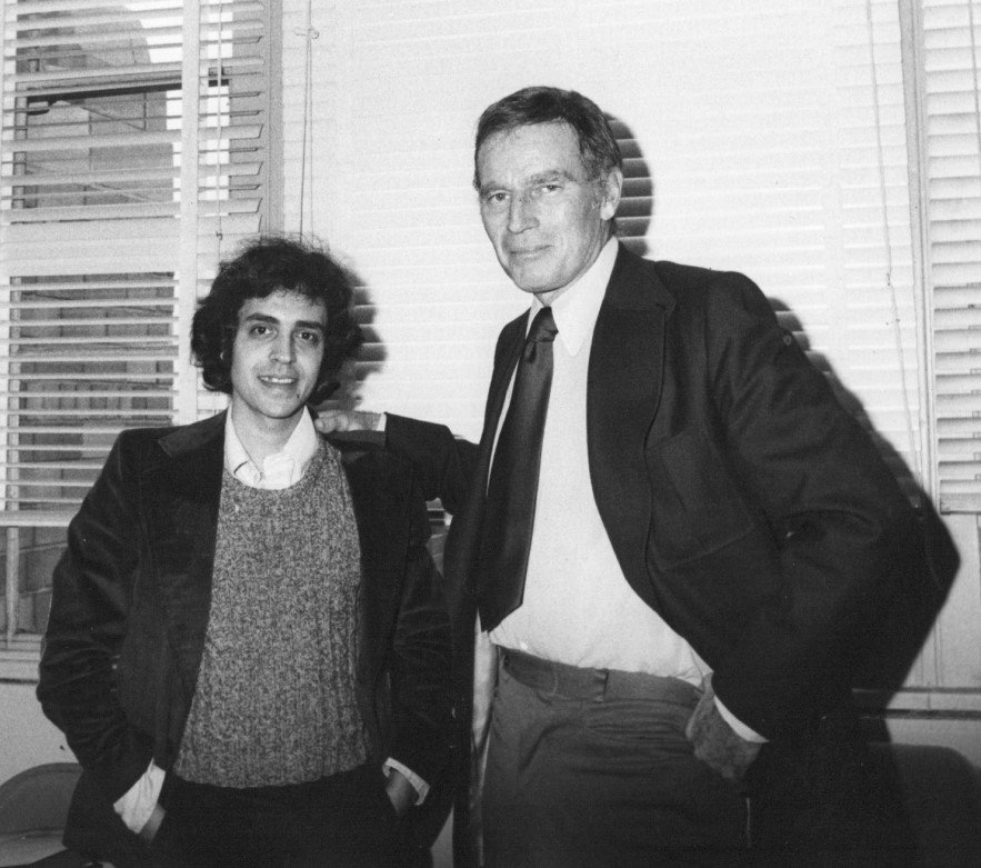 Two men wearing blazers pose in front of a window