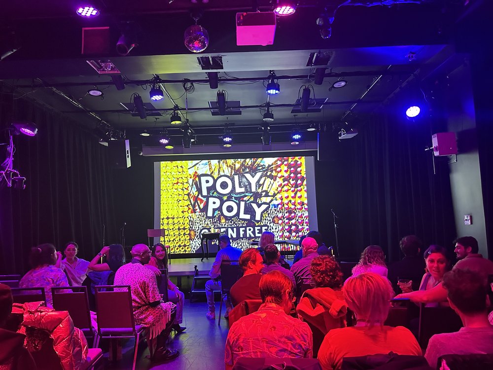 A group of people sit in a theater looking at a sign that says "Poly Poly Oxen Free."