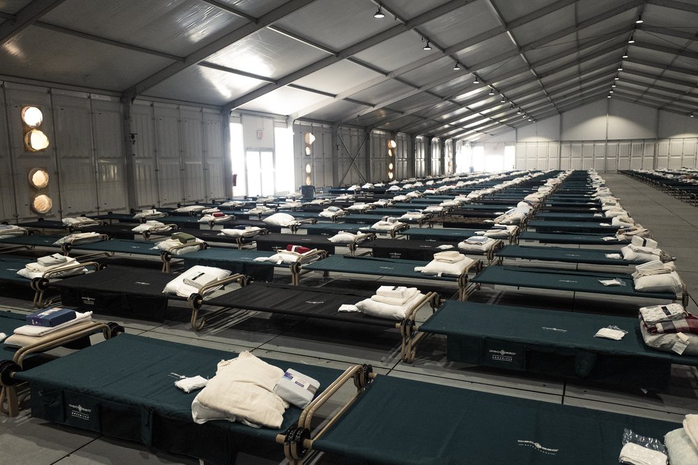 Rows of green and black cots inside a large, white-paneled building.