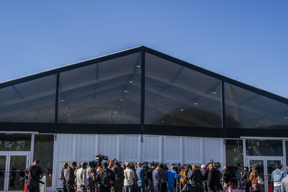 A large huddle of press members, many news cameras, outside a large white-paneled building with a black roof.