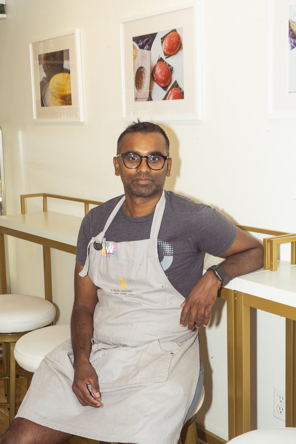 A person wearing an apron sit on a stool, beneath framed pictures on a wall.