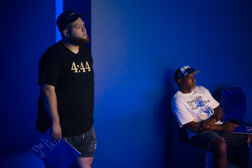 A person wearing a black t-shirt that reads "4:44" stands looking at a screen in blue room.