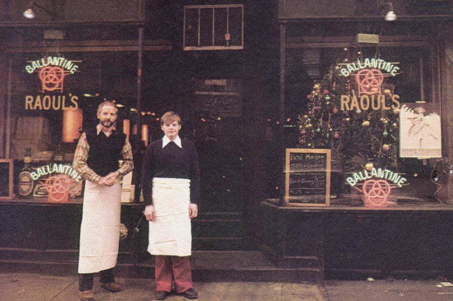 Two men stand in front of a window.