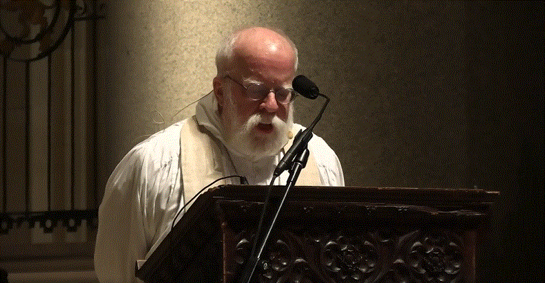 An older man dressed in religious clothing speaks at a podium.
