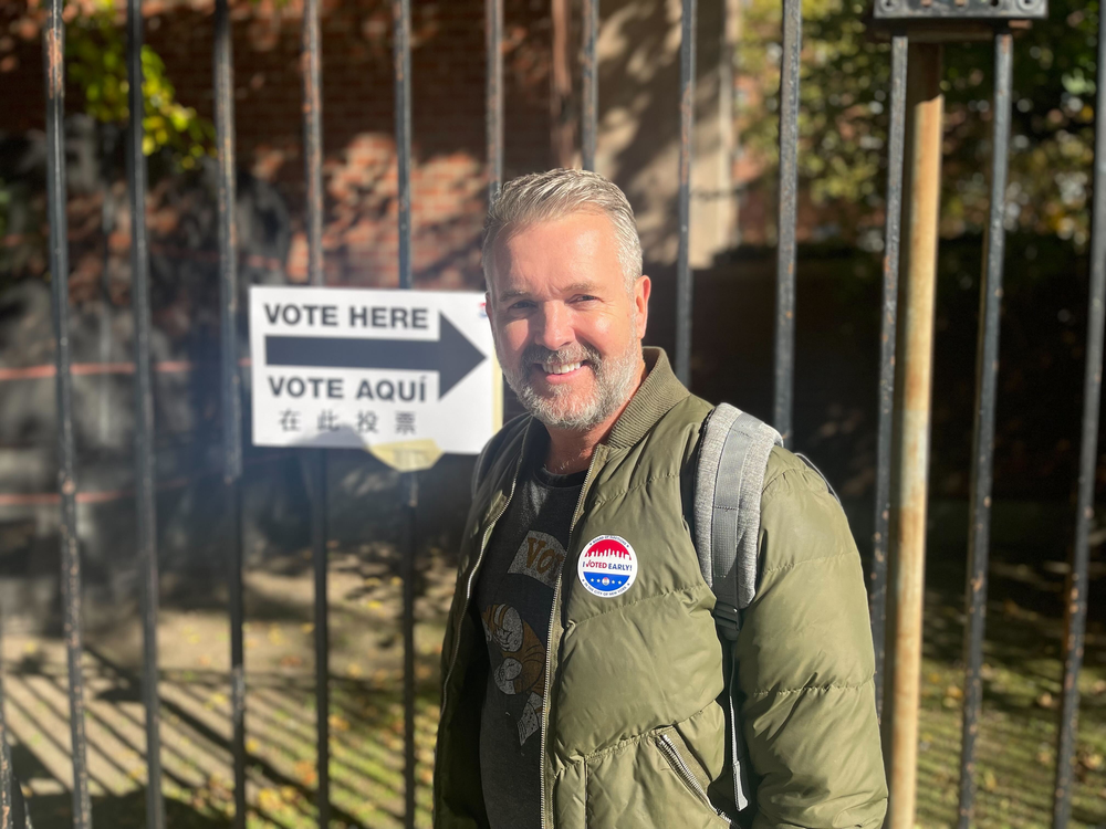 A man in a jacket standing outdoors with a voting sign behind him.